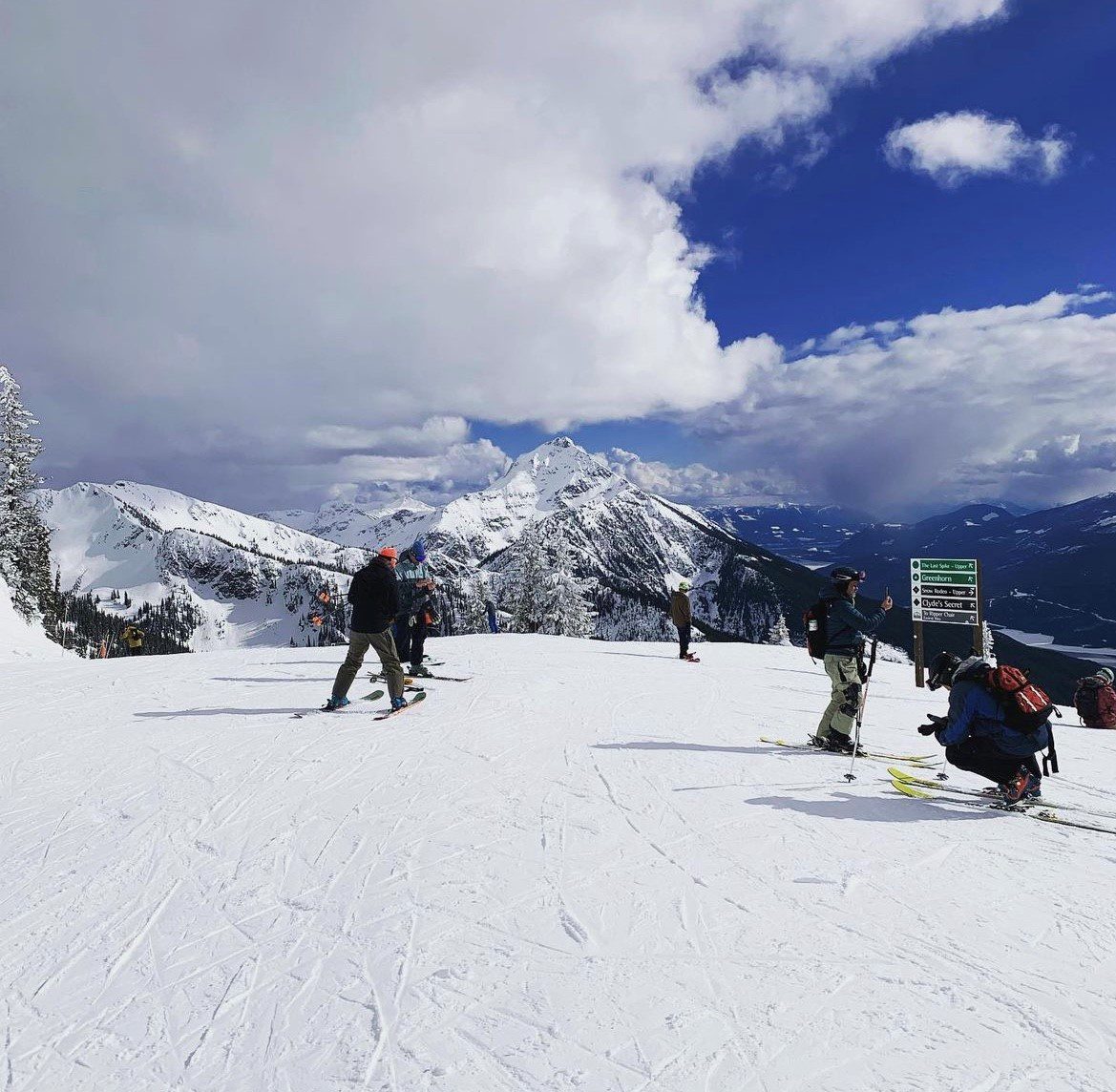 sitepic1 Several skiers and snowboarders on a snow-covered mountain with cloudy sky and distant peaks in the background.