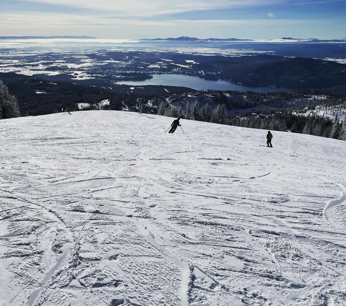 sitepic11 Three people skiing down a snowy slope with a panoramic view of lakes, forests, and distant mountains under a clear blue sky.