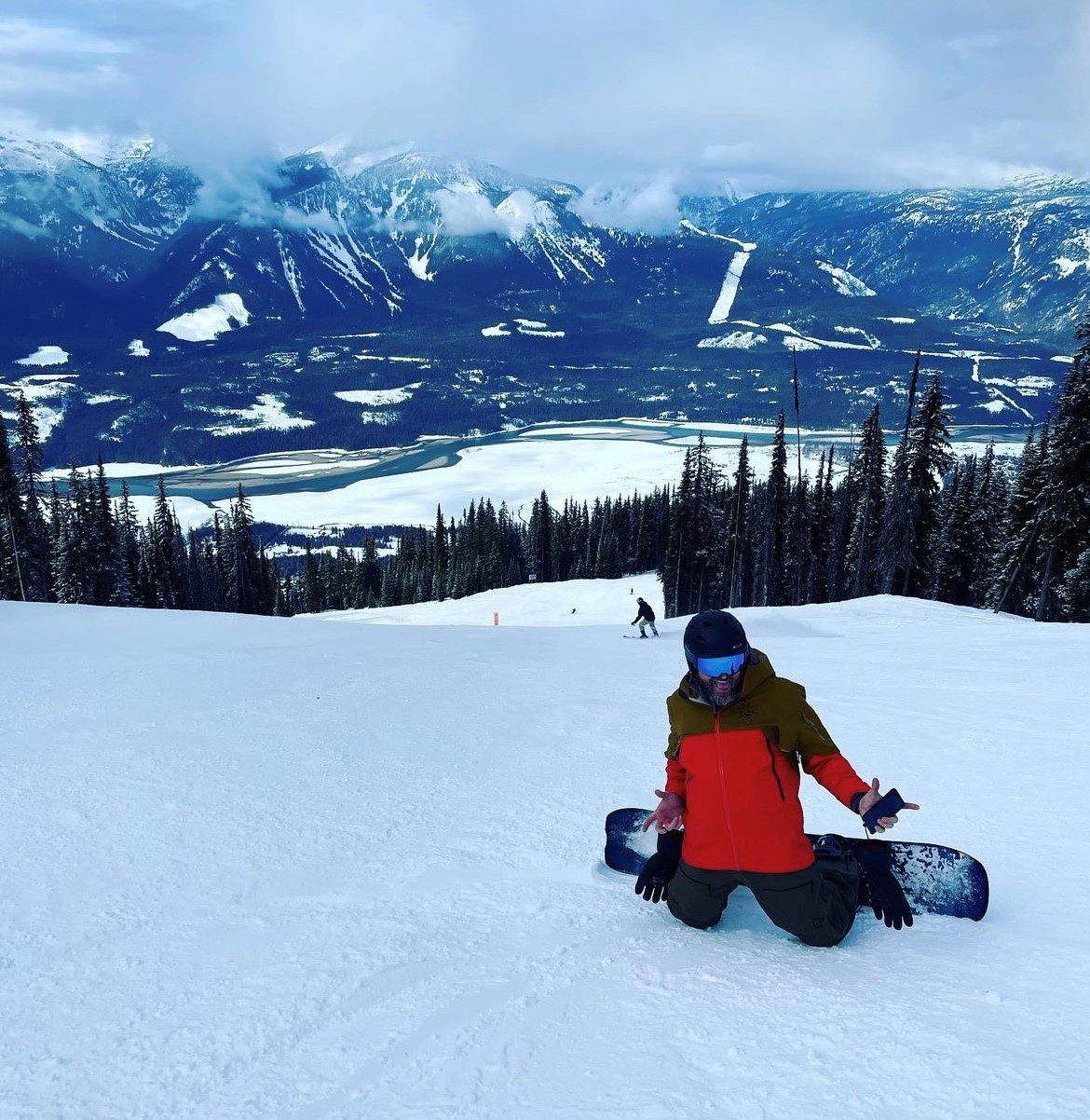 sitepic4 Snowboarder kneeling on a snowy slope with a scenic mountain and forest backdrop, wearing a red and green jacket and helmet. Another person is snowboarding downhill in the distance.