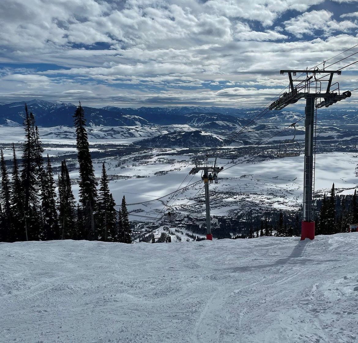 sitepic6 Snow-covered landscape with a ski lift and distant town in the valley. Tall trees on the slope, mountains visible on the horizon under a partly cloudy sky.